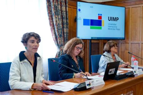 Rosa Martínez, secretaria de Estado de Derechos Sociales, participando esta semana en los cursos de verano de la Universidad Internacional Menéndez Pelayo en Santander, sentada junto a dos personas a una mesa, con documentos, micrófonos y una pantalla grande de fondo.